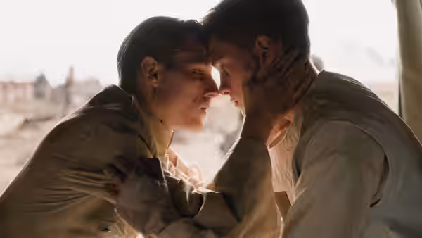 Two young men lean close together in a softly lit room