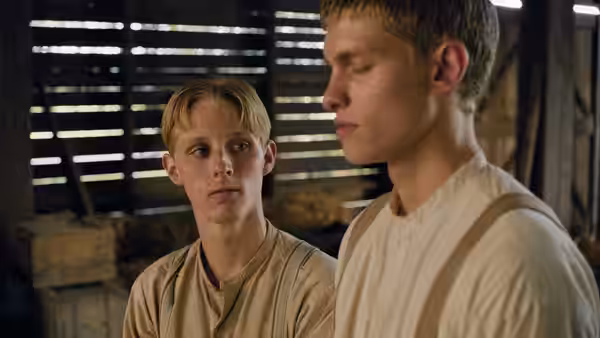 Two young men stand together inside a dim wooden barn