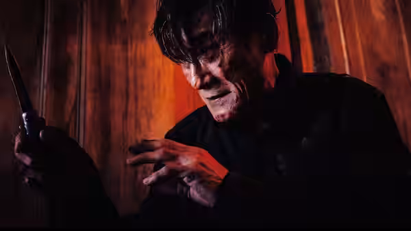 A bloodied man grips a knife in a dim wooden room