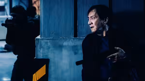 Wong Tak Chung (Jackie Chan) takes cover behind a pillar as an armed figure aims nearby