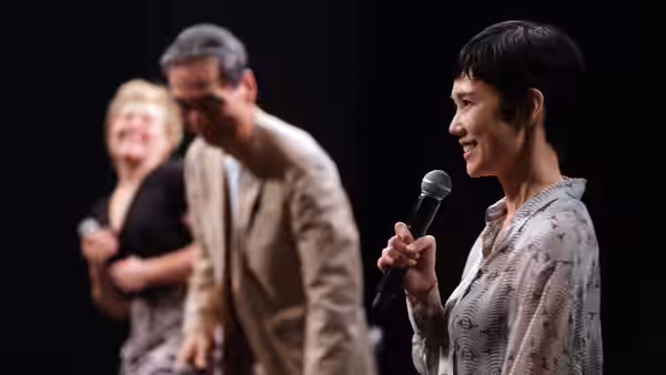 Mari Morisaki (Tao Okamoto) speaks into a microphone onstage as two people stand behind her