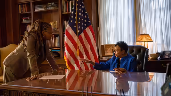 Two women speak across a polished desk, one gesturing beside an American flag