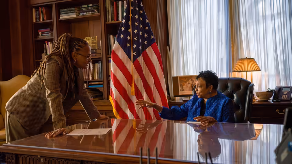 Two women speak across a polished desk, one gesturing beside an American flag