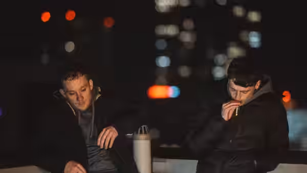 Two young men sit by a railing at night, one smoking beside a takeaway cup