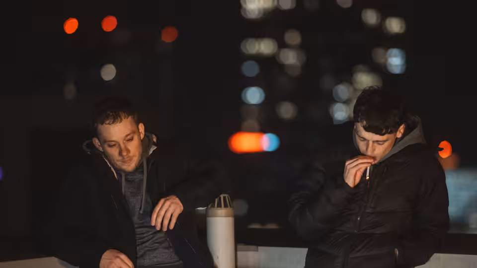 Two young men sit by a railing at night, one smoking beside a takeaway cup
