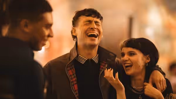 A laughing young man and woman stand arm in arm as another man watches