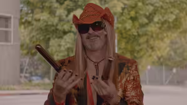 A man in a red cowboy hat and long blond wig holds a wooden object