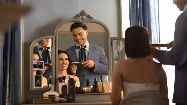 A man helps a seated woman with her makeup as she gets ready in a dressing room