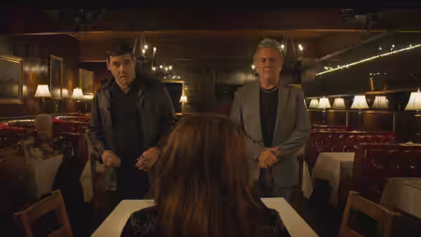 Two men address a woman seated across from them in a dim restaurant