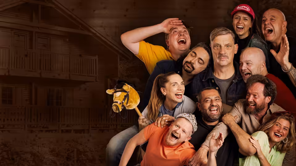 A group of comedians crowd together, laughing and posing before a wooden auditorium backdrop
