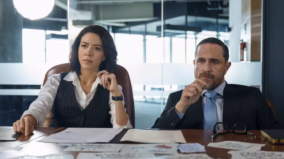 A woman and a bearded man sit at a conference table reviewing documents