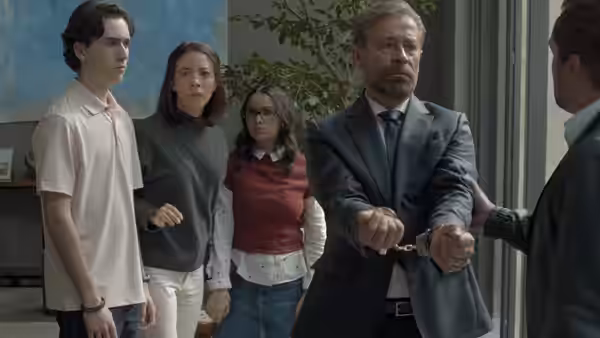 An older man stands handcuffed beside three young people as another man holds his arm