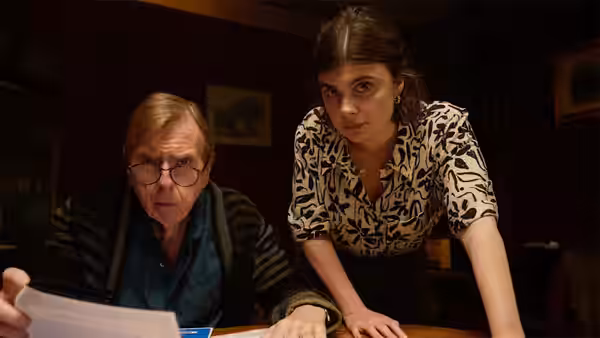 John Chapel (Timothy Spall) and Janie Mallowan (Gwyneth Keyworth) study papers at a table