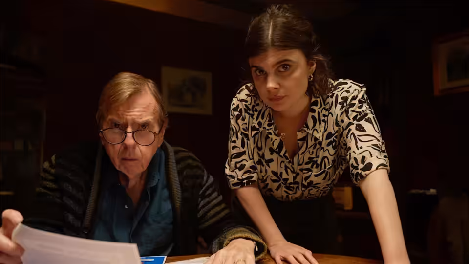 John Chapel (Timothy Spall) and Janie Mallowan (Gwyneth Keyworth) study papers at a table