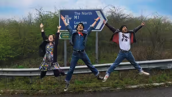 Three young people leap beside a motorway sign pointing toward London