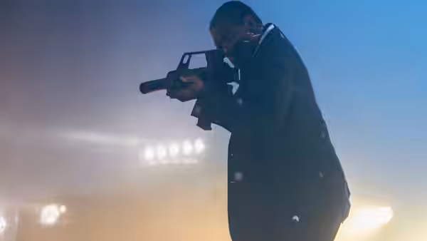 A suited man aims a rifle beneath bright outdoor lights