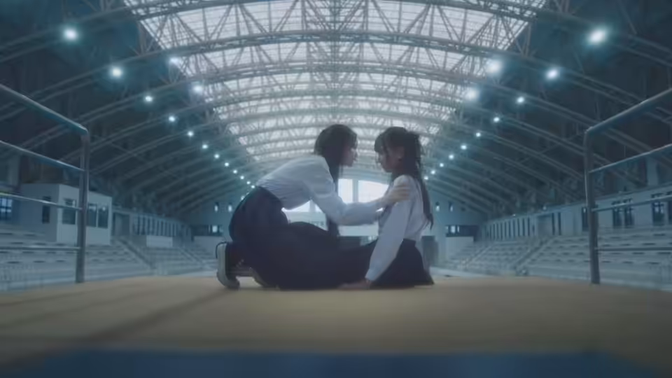 Two young women in school uniforms kneel facing each other on an indoor sports court