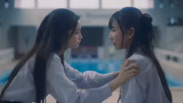 Two schoolgirls face each other beside an indoor swimming pool, holding each other by the shoulders