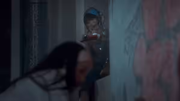 A helmeted woman points a red spray can toward another woman in a dim corridor