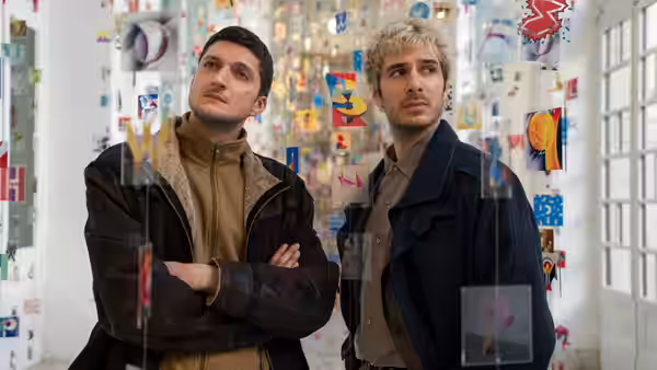 Two men examine colorful artworks displayed on glass walls in a gallery