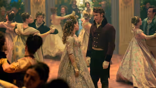 A woman in a patterned gown faces a uniformed man as couples dance in a ballroom