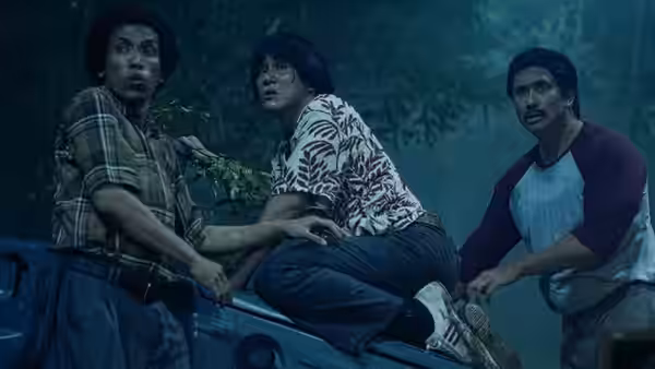 Three men crouch together on a vehicle beneath trees, looking alarmed