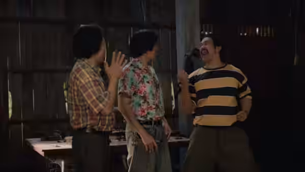Two men face a gesturing man inside a shadowy wooden room