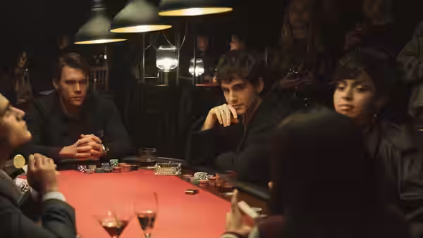 Young people sit around a poker table beneath hanging lamps