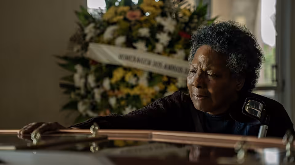Vicentina (Rejane Faria) leans over a coffin beneath a funeral wreath