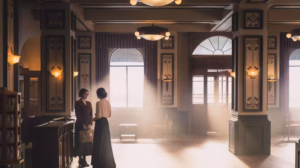 Two women stand beside the reception desk in a sunlit hotel lobby