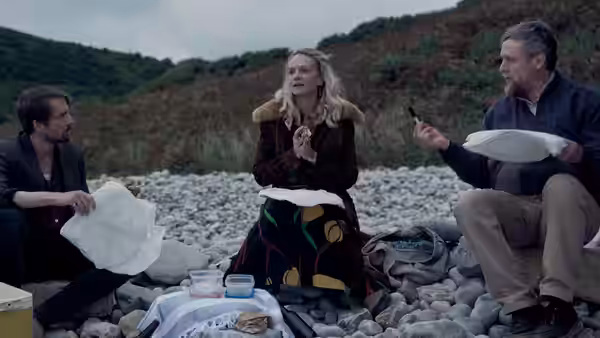 Three people share a picnic on a stony beach beside the sea