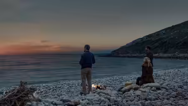 Three figures gather around a small fire on a beach at dusk