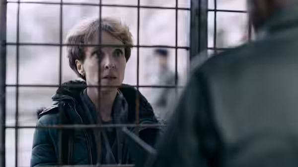 A short-haired woman speaks through a gridded glass partition to someone nearby