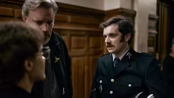 A mustached uniformed officer speaks with two men in a wood-paneled room