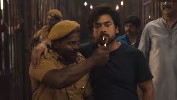 A uniformed officer lights a cigarette in a curly-haired man's mouth in a crowded corridor
