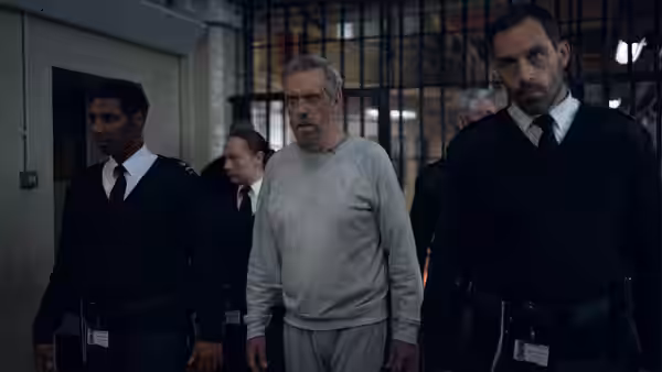 Felix Carmichael (Hugh Laurie) is escorted by guards through a barred facility