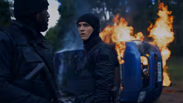 Two men in tactical gear stand beside a burning vehicle