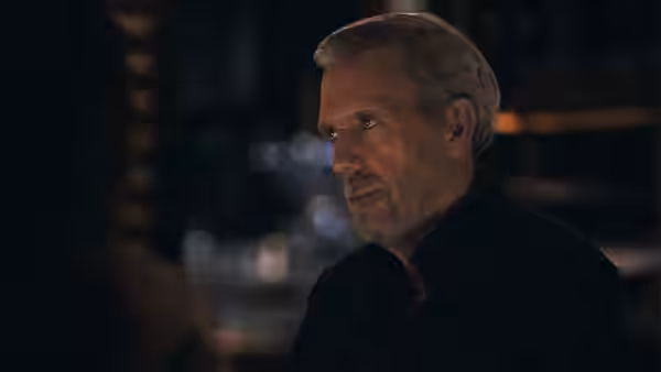 John Carmichael (Stephen Dillane) sits in a dim room, facing someone across the table