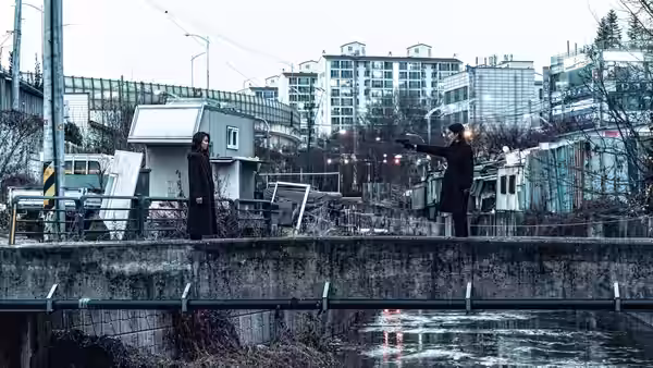 Two women face each other across a narrow stream in an urban neighborhood