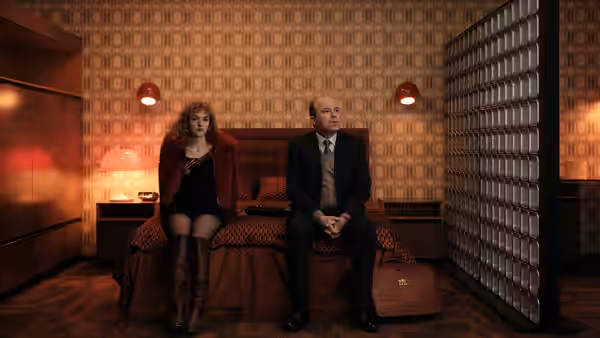 A woman and a suited man sit apart on a patterned sofa in a warmly lit room