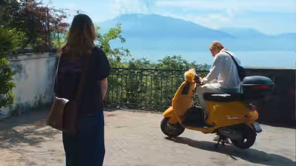 A young woman watches a man on a yellow scooter beside a lake