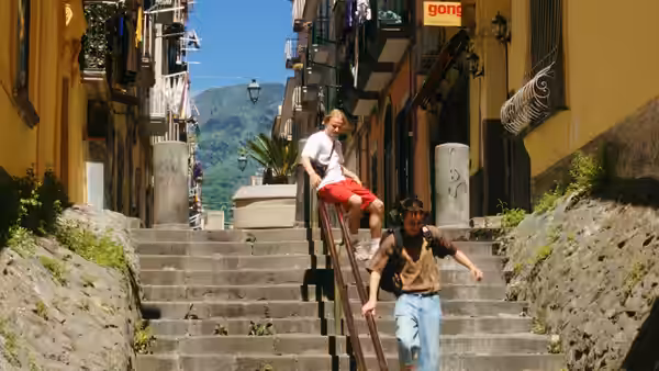 A young man descends a steep stone staircase as another sits on the railing above