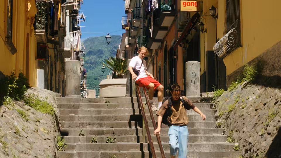 A young man descends a steep stone staircase as another sits on the railing above