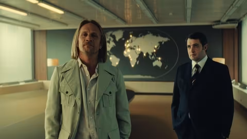 Arnaud de Toledo (Jérémie Renier) stands beside a suited man in a modern office lobby