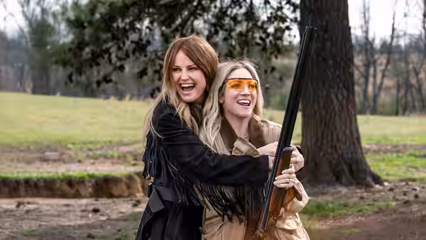 A woman embraces a younger woman holding a rifle in a grassy clearing