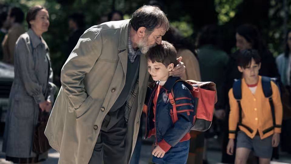 An older man kisses a schoolboy goodbye beside other children with backpacks