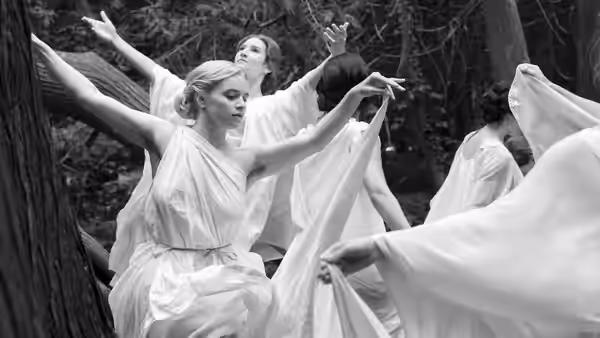 Women in flowing white dresses extend their arms in a woodland dance