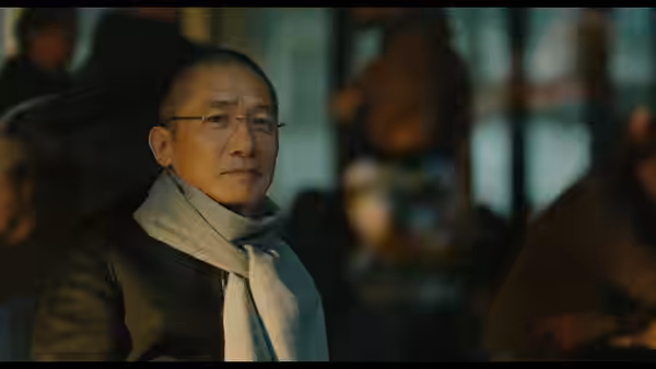 Tony Leung sits among other people indoors, wearing glasses and a scarf