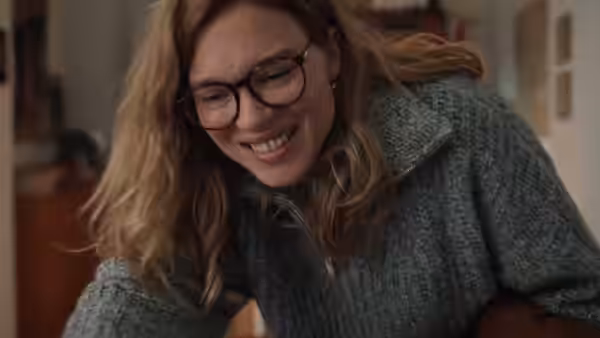 A bespectacled woman smiles as she leans forward indoors