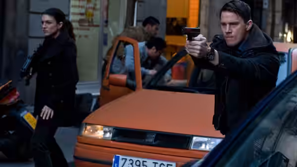Mallory Kane (Gina Carano) holding a gun beside an armed man by an orange car